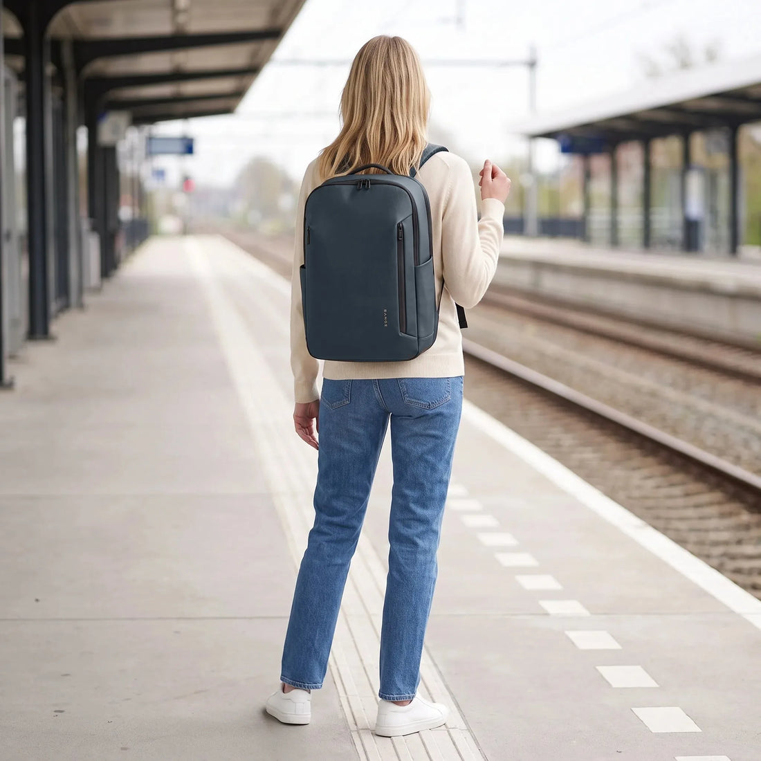 Vrouw draagt de donkerblauwe DS Shield laptoprugzak op een treinperron, klaar voor vertrek