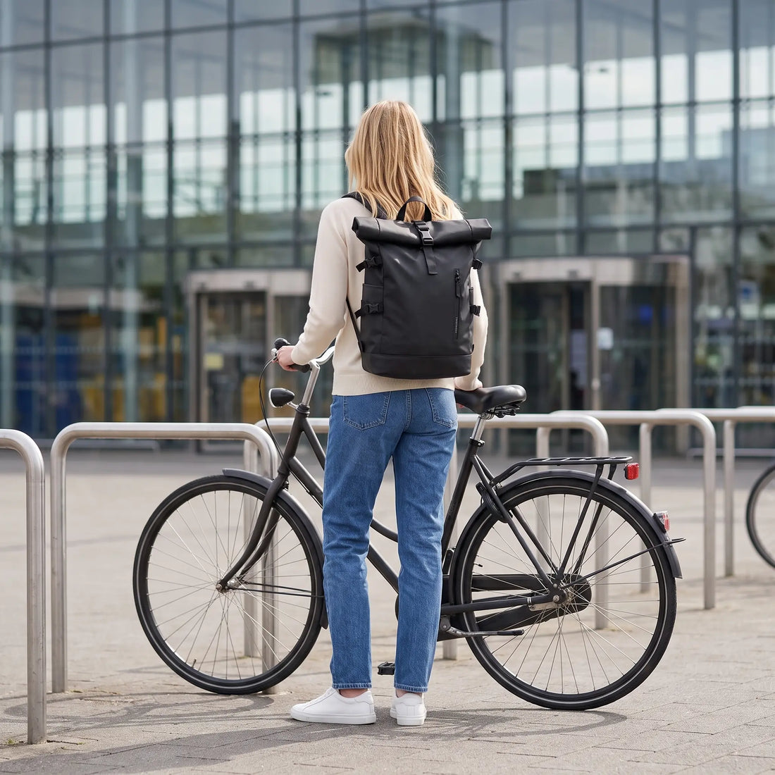 Vrouw met de zwarte DS Elevate rolltop rugzak staat met stadsfiets voor een stationsgebouw