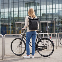 Vrouw met de zwarte DS Elevate rolltop rugzak staat met stadsfiets voor een stationsgebouw